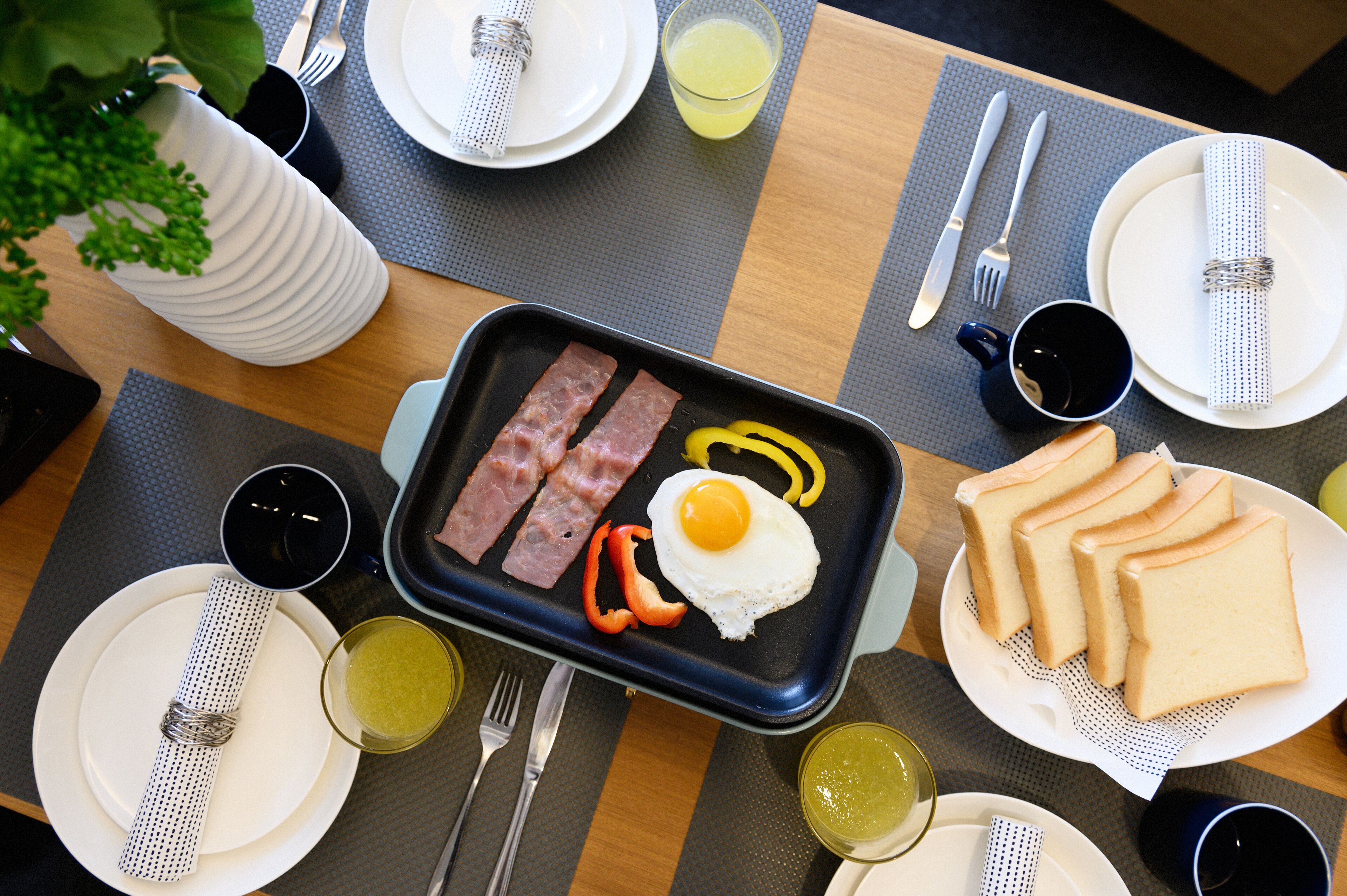 A breakfast tray with bacon, egg, and peppers on a black plate, surrounded by plates, cups, and cutlery on a table.