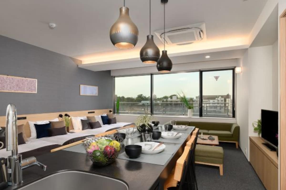 A modern hotel room with a dining area, a sofa, a TV, and a large window offering a city view.