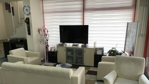 Uma sala de estar com sofá, poltronas, mesa de centro de vidro, televisão e um armário.