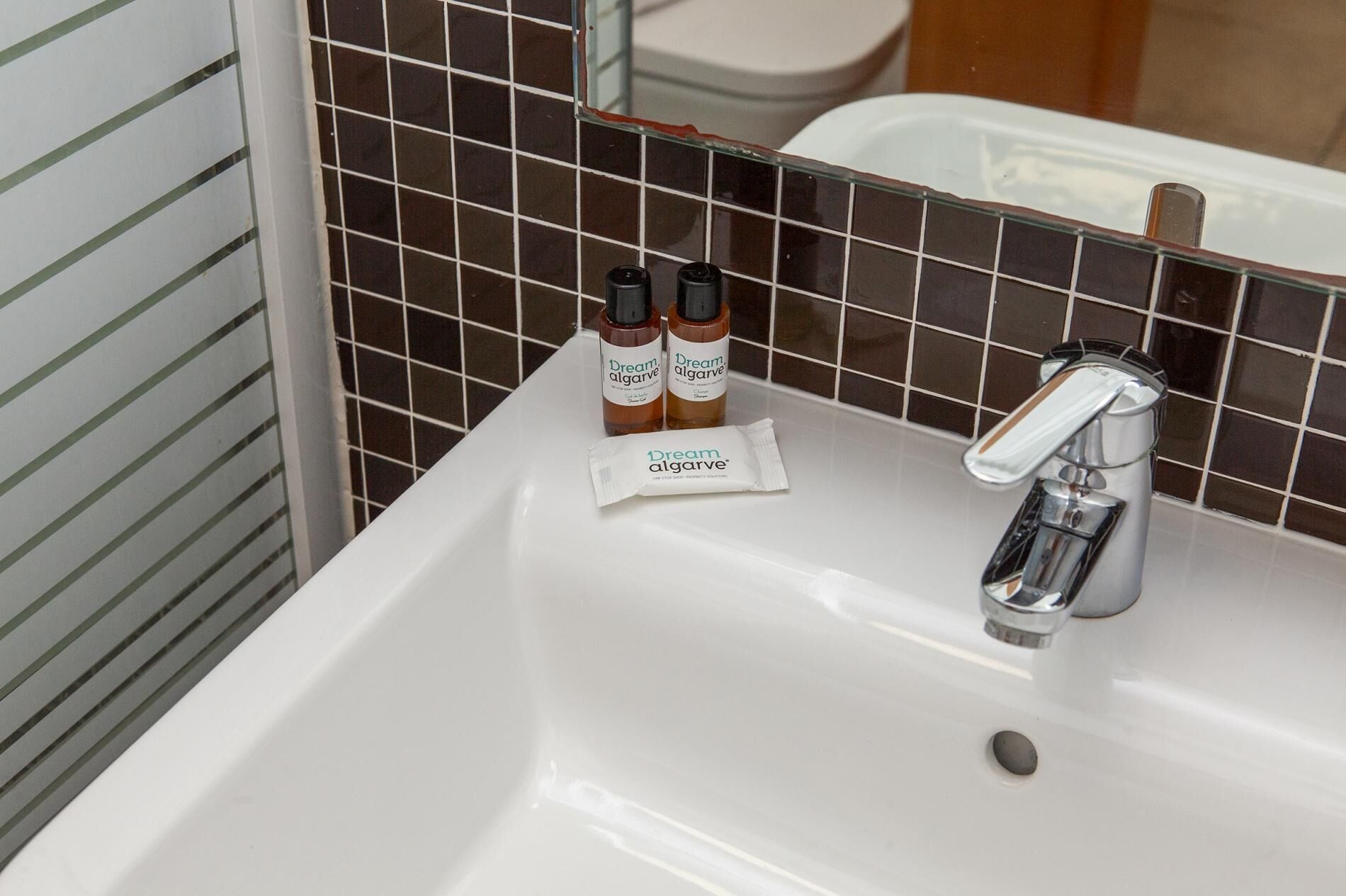 Uma pia de banheiro com torneira cromada, duas garrafas de shampoo Dream Algarve e um sachê do mesmo produto.