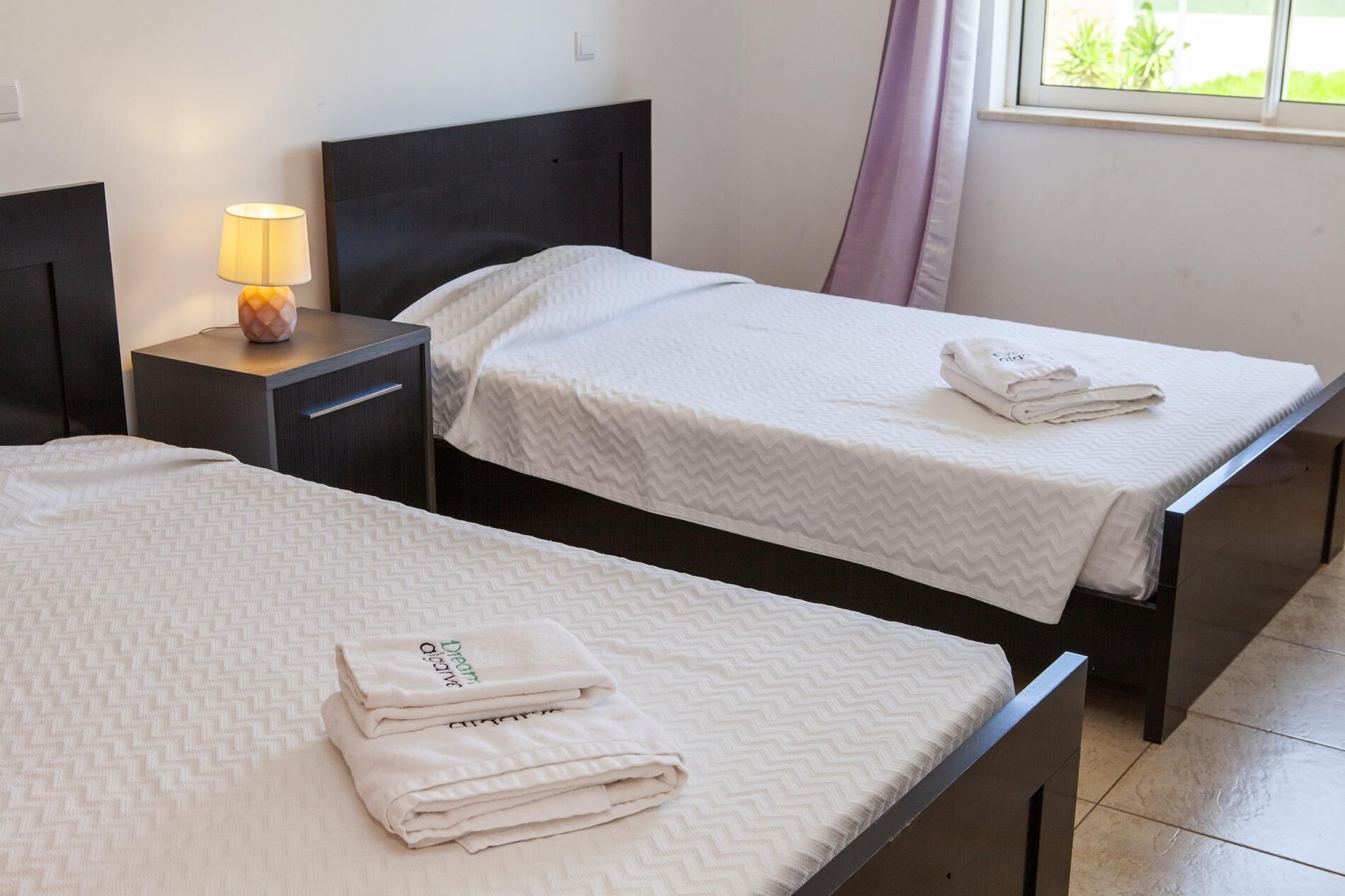 Two single beds with white bedding and folded towels, a bedside table with a lamp, and a window with curtains.