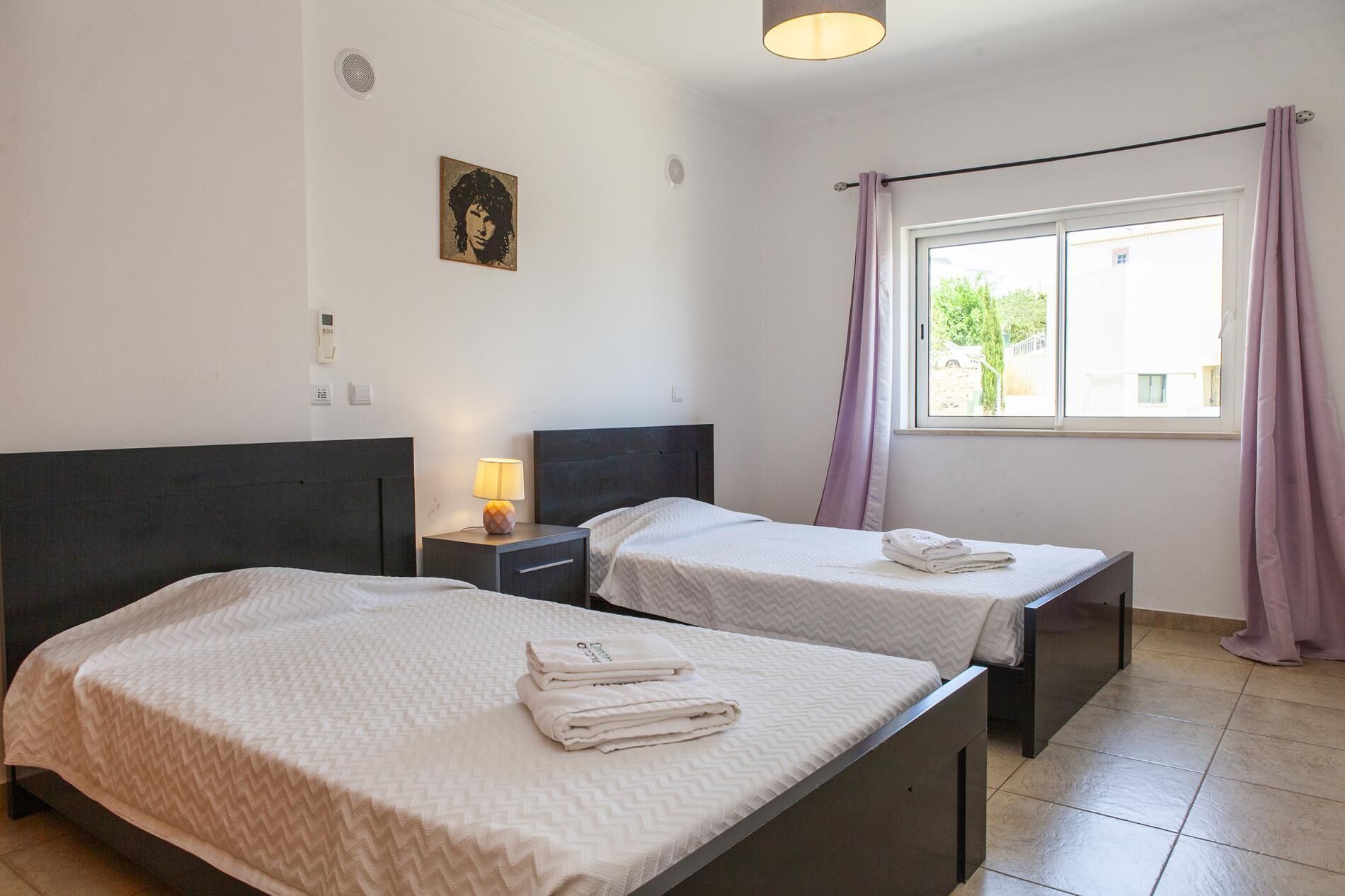A hotel room with two beds, a bedside table with a lamp, a window with curtains, and a wall-mounted light switch.