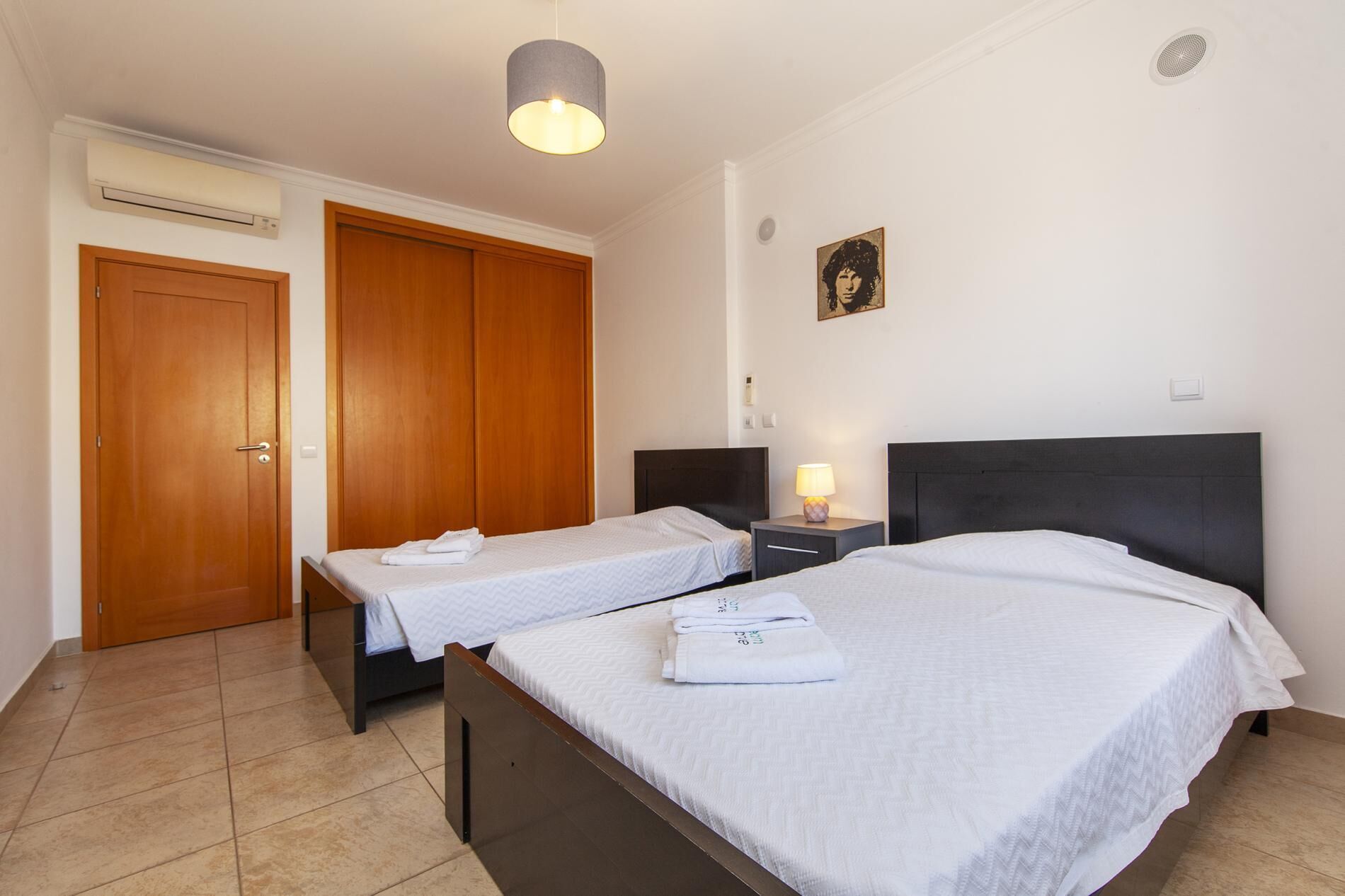 Una habitación de hotel con dos camas, un armario de madera, una mesita de noche con lámpara y un equipo de aire acondicionado.