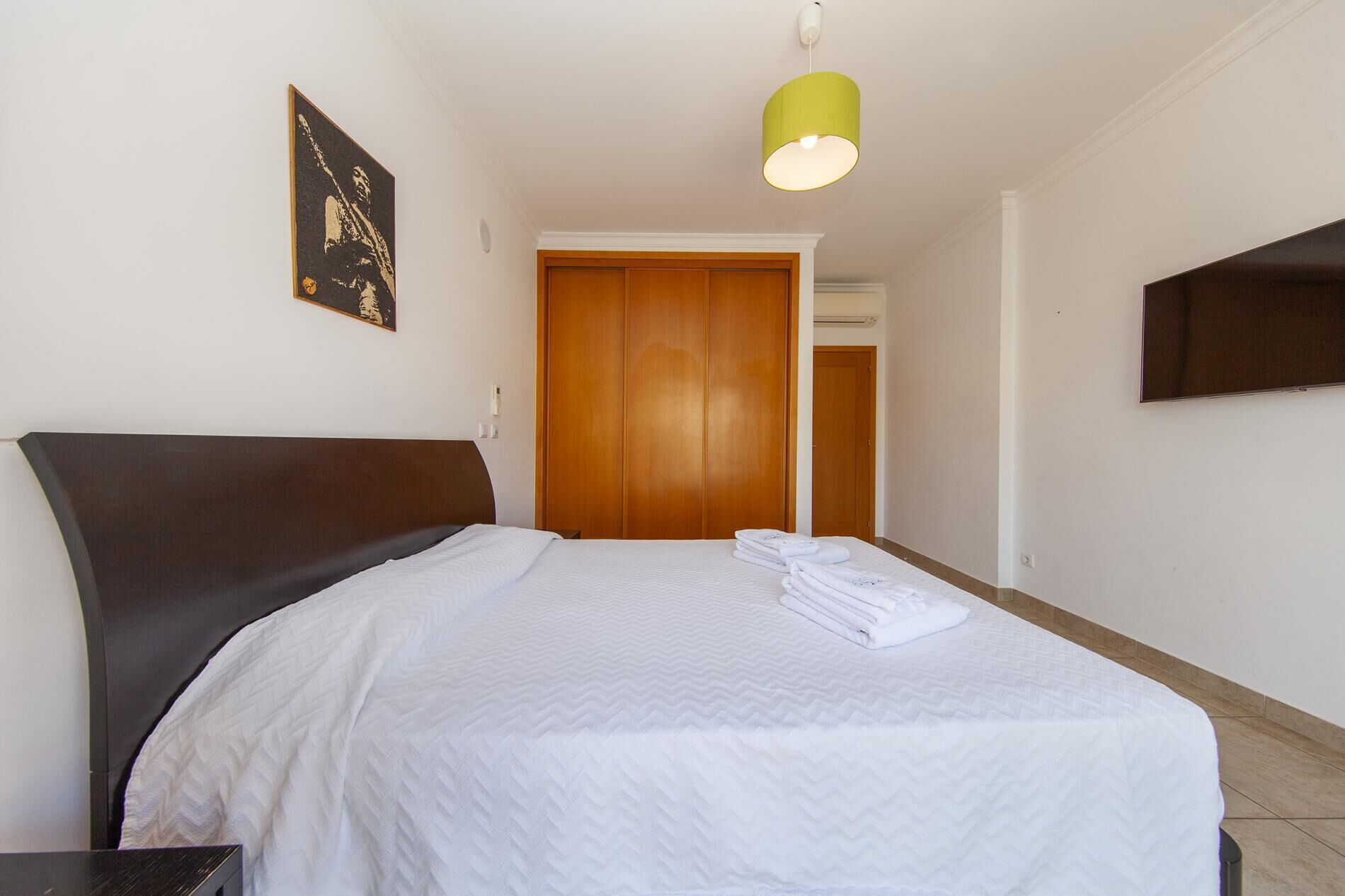 A neatly made bed with white bedding, a wooden headboard, and a yellow pendant light.