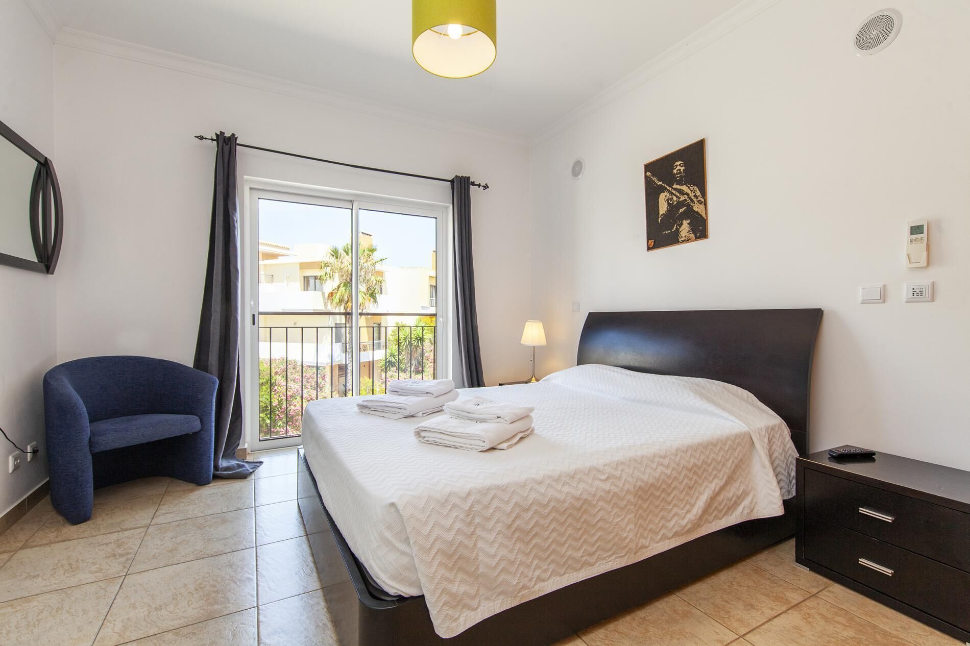 Une chambre d’hôtel avec un lit, une chaise, une table de chevet, une lampe et une fenêtre avec des rideaux.
