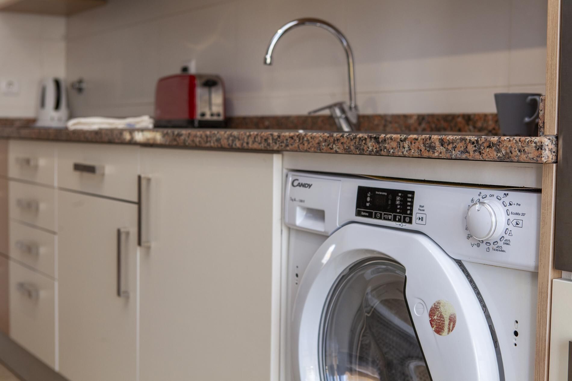 A front-loading washing machine with a control panel, a granite countertop, a red toaster, and a stainless steel faucet.
