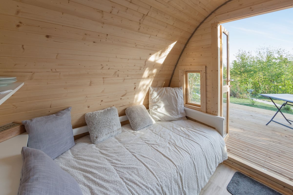 A cozy, wooden interior with a curved ceiling, a bed with pillows, and a view of a deck and greenery outside.