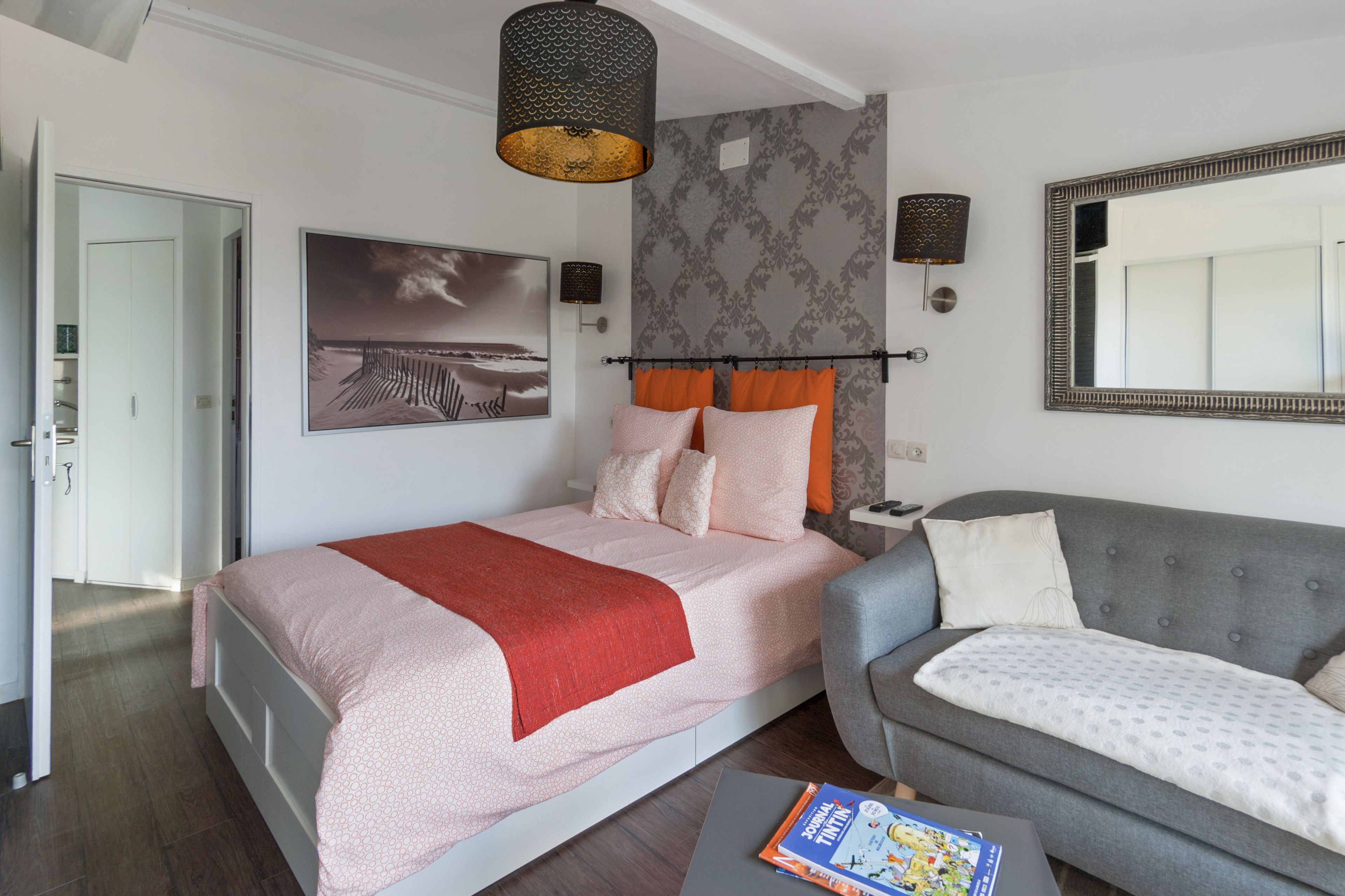 A modern bedroom with a bed, a grey sofa, a large mirror, and a painting on the wall.