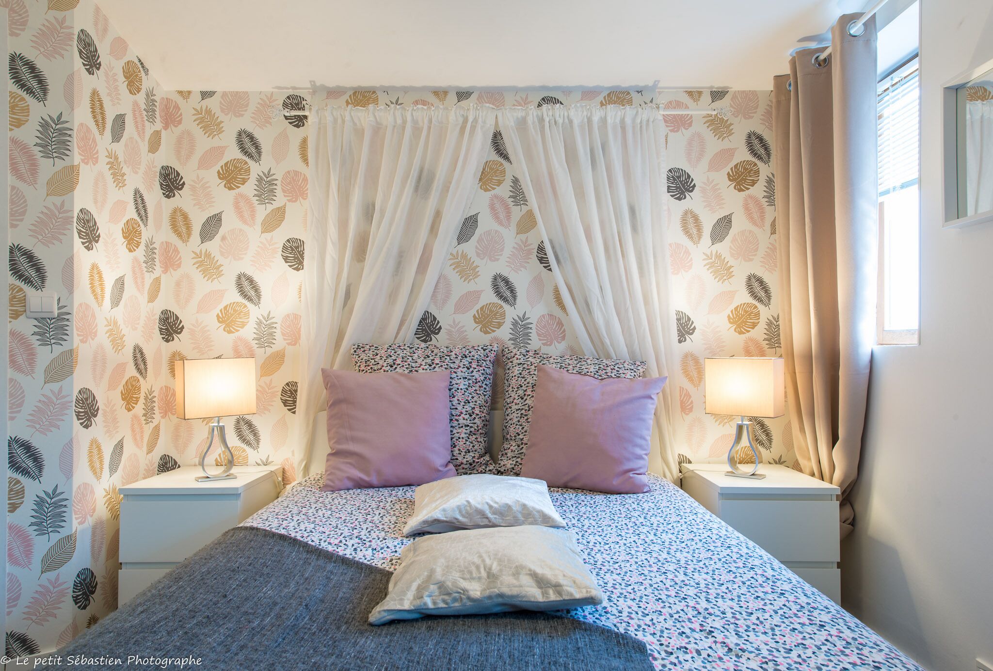 Une chambre à coucher avec un lit à baldaquin, un papier peint à motifs floraux, deux lampes de chevet et une fenêtre avec des rideaux.