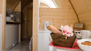 Un interior rústico de madera con ventana en marco de madera, una zona de cocina con armarios y una cafetera, y una mesa con una cesta de pan y una flor.