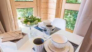 Interior de una cabaña de madera con una mesa de comedor dispuesta para dos personas, un jarrón con flores, dos tazas y un plato.