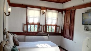 A bedroom with a wooden ceiling, two windows with curtains, a bed with pillows, a radiator, and a television on the wall.