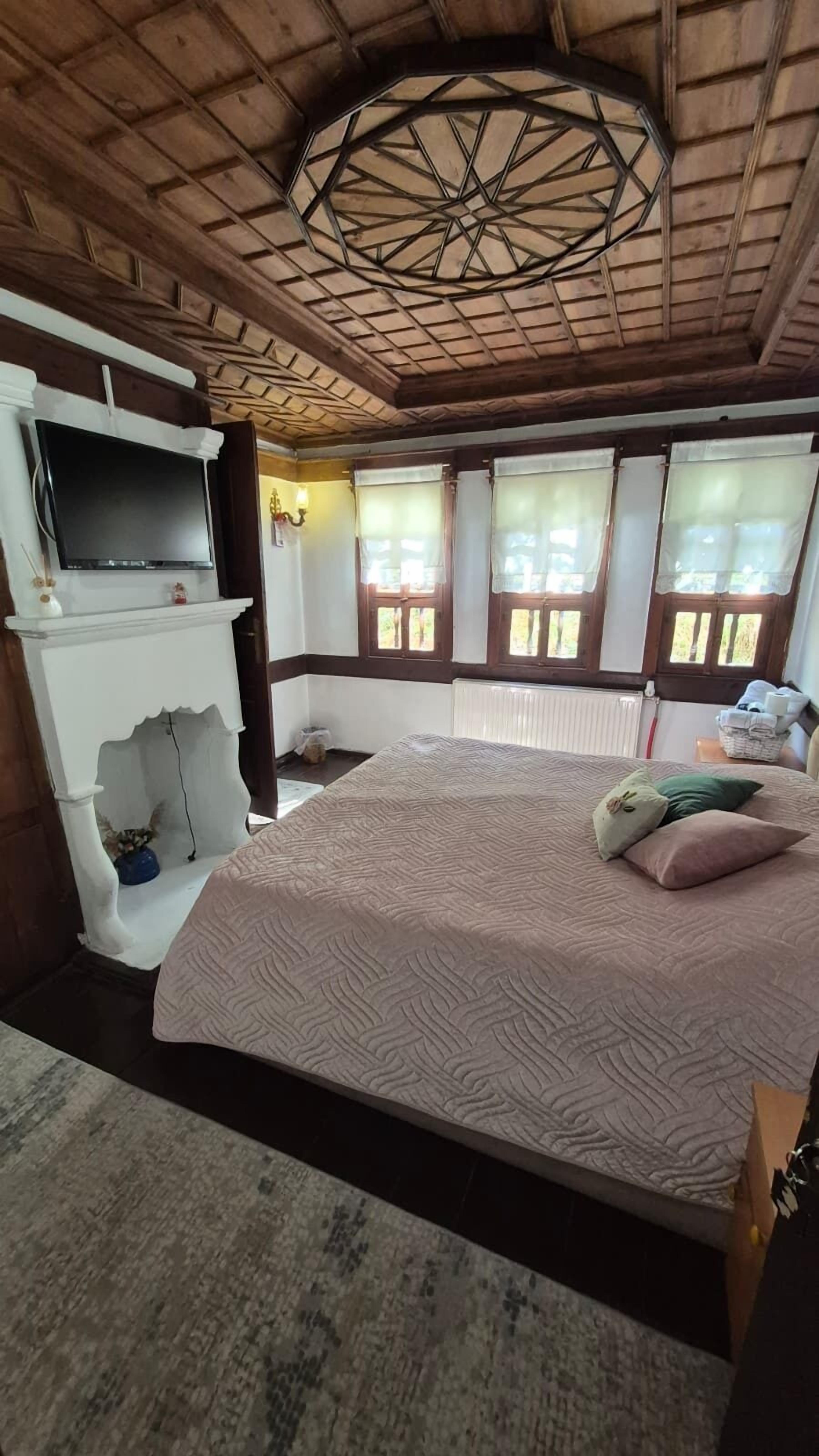 A bedroom with a large bed, a TV mounted on a white wall, a fireplace, and windows with curtains.