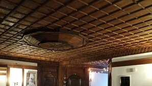 A room with a wooden ceiling, a large red patterned rug, a bed with a white bedspread, and a small table with a decorative item.