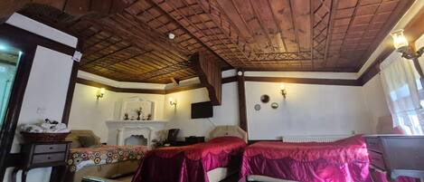 A room with wooden ceiling beams, two beds with red covers, a fireplace, and a television mounted on the wall.