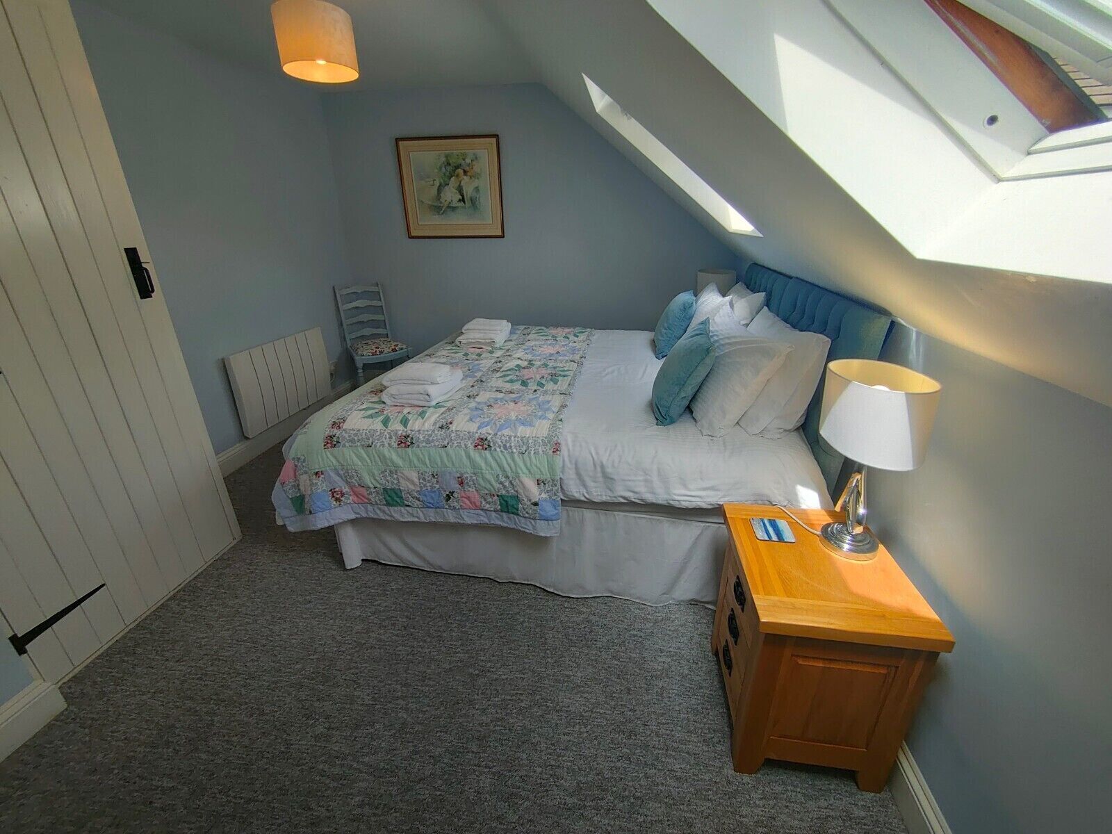 A bedroom with a bed, a bedside table with a lamp, a radiator, a window, and a picture on the wall.
