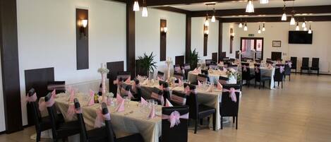 Un salón de banquetes con varias mesas dispuestas para un evento formal, con sillas negras adornadas con cintas rosas, manteles blancos y lámparas colgantes.