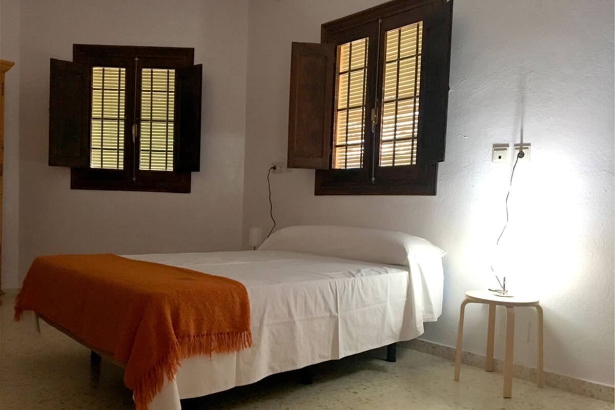 A bedroom with a bed, an orange throw blanket, two windows with wooden shutters, and a bedside table with a lamp.