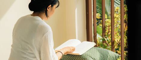 A person sitting on a bench reading a book in a well-lit room with a view of greenery outside.