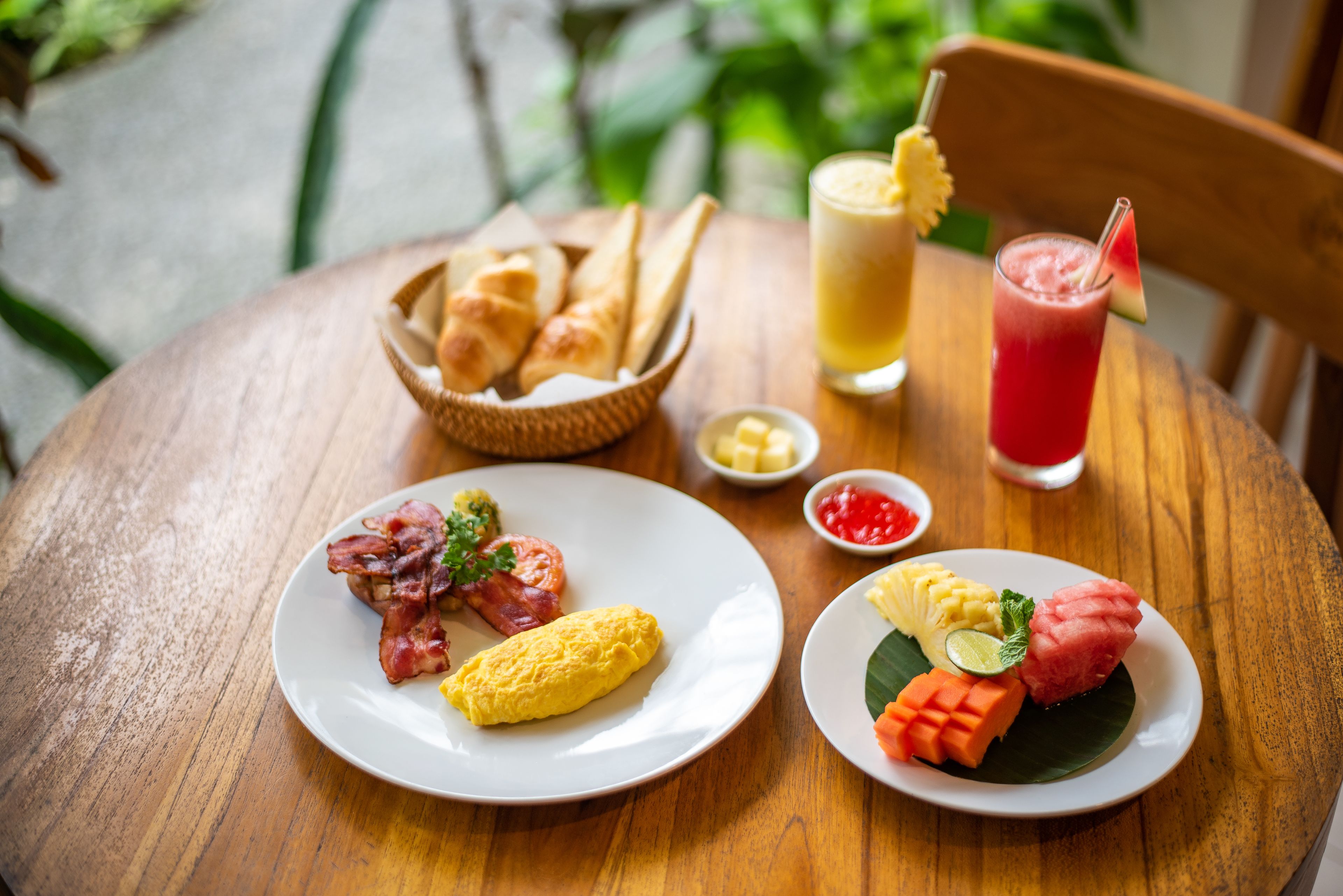 A wooden table with plates of food, including eggs, bacon, and tropical fruits, and a drink with a straw.