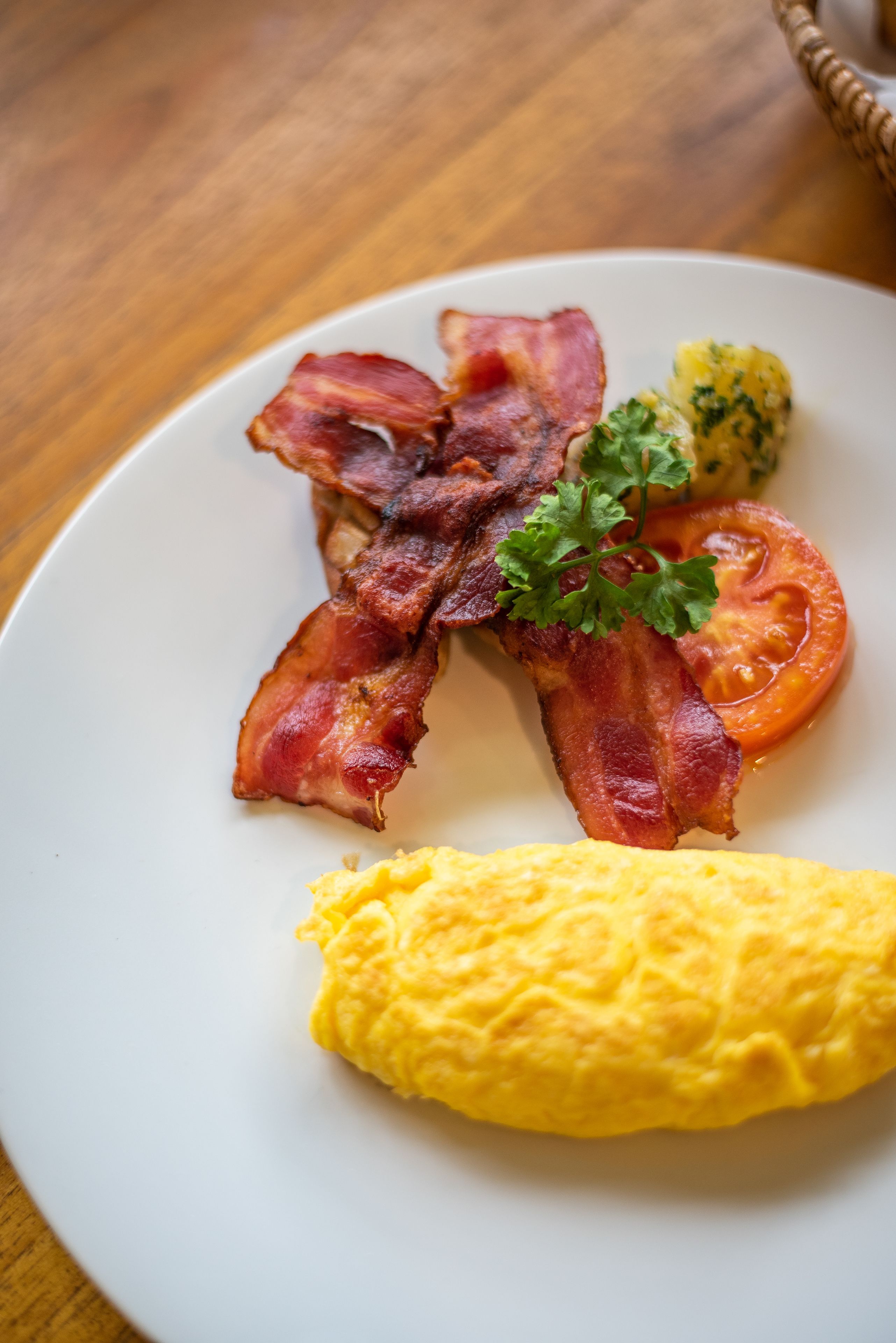A plate with bacon, a tomato slice, scrambled eggs, and a garnish on a wooden table.