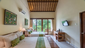 A hotel room with two beds, a desk, a TV, and a balcony with a view of greenery.