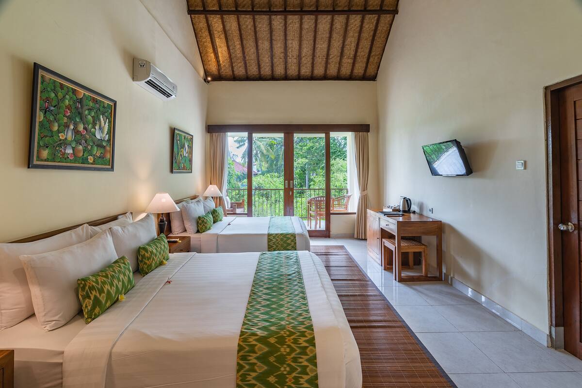 A hotel room with two beds, a desk, a TV, and a balcony with a view of greenery.