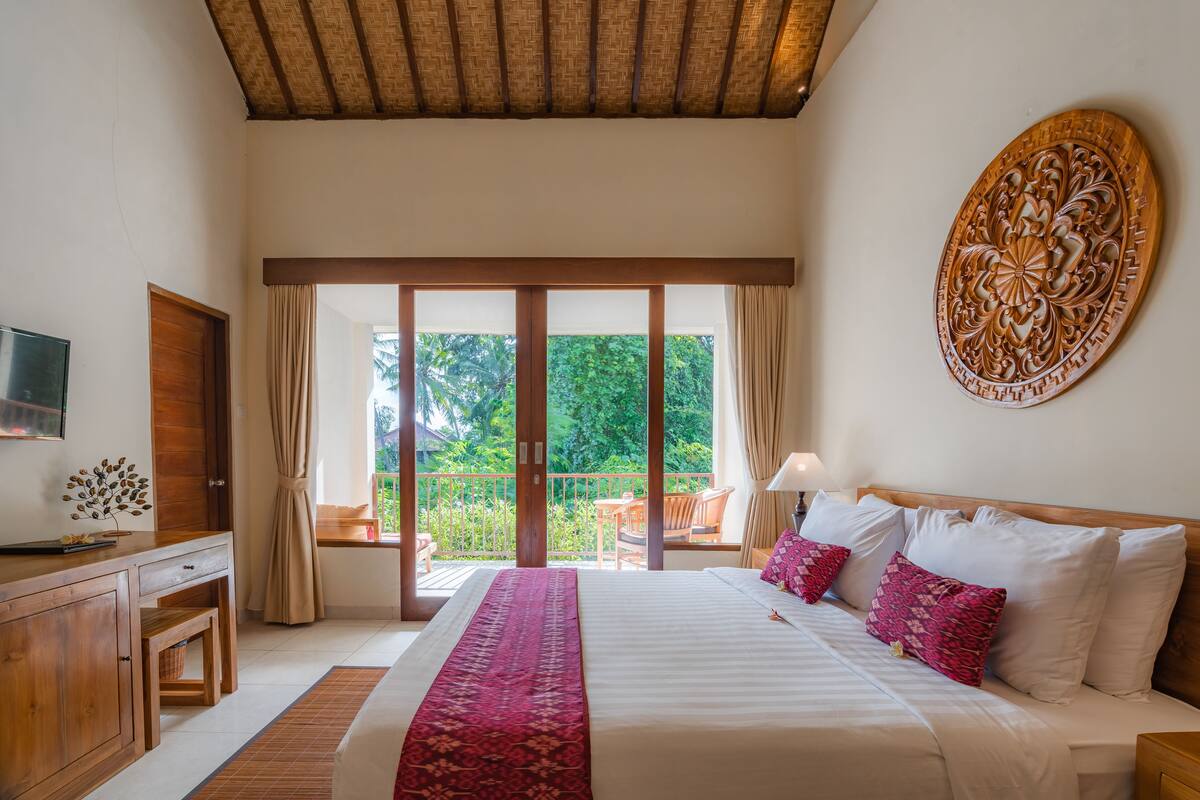 A bedroom with a large bed, a wooden desk, a television, and a balcony with a view of greenery.