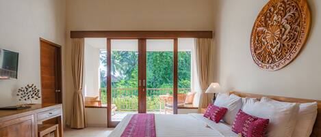 A bedroom with a large bed, a wooden desk, a television, and a balcony with a view of greenery.