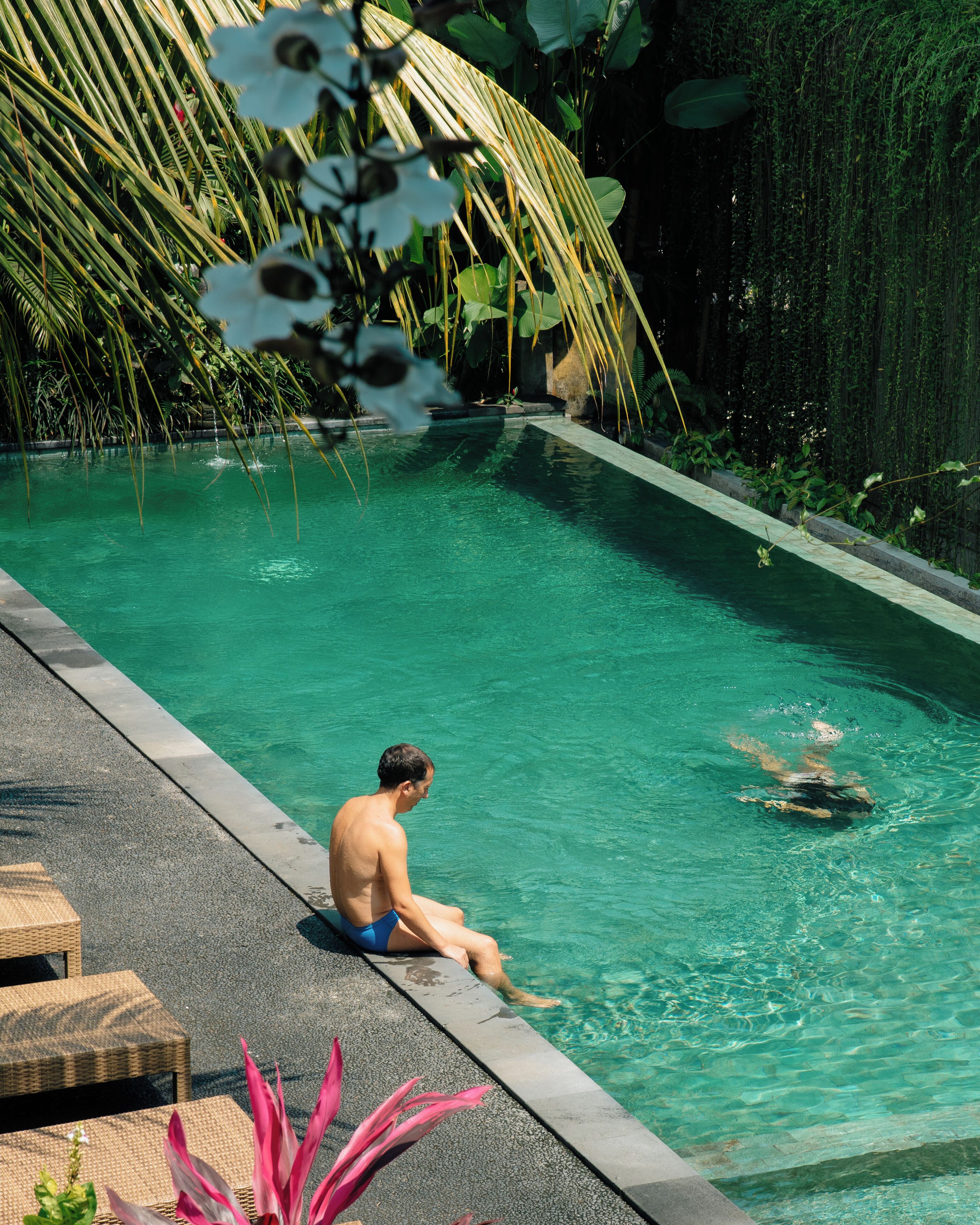 A man sitting by a pool with clear water, surrounded by lush greenery and a stone deck.