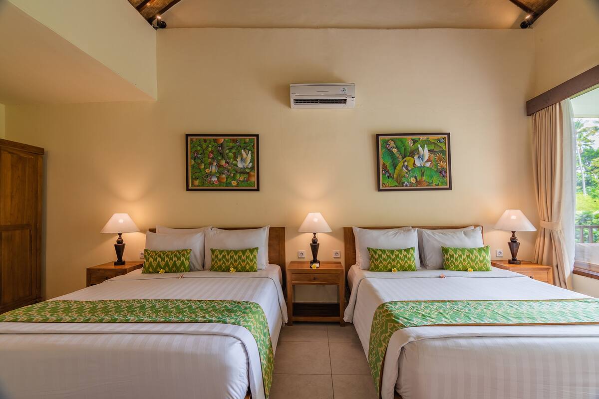 A hotel room with two beds, each with a green patterned bedspread, two wall-mounted air conditioners, and two framed floral paintings.