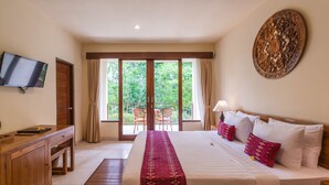 A hotel room with a bed, a TV, a wooden desk, a balcony with chairs, and a view of greenery.