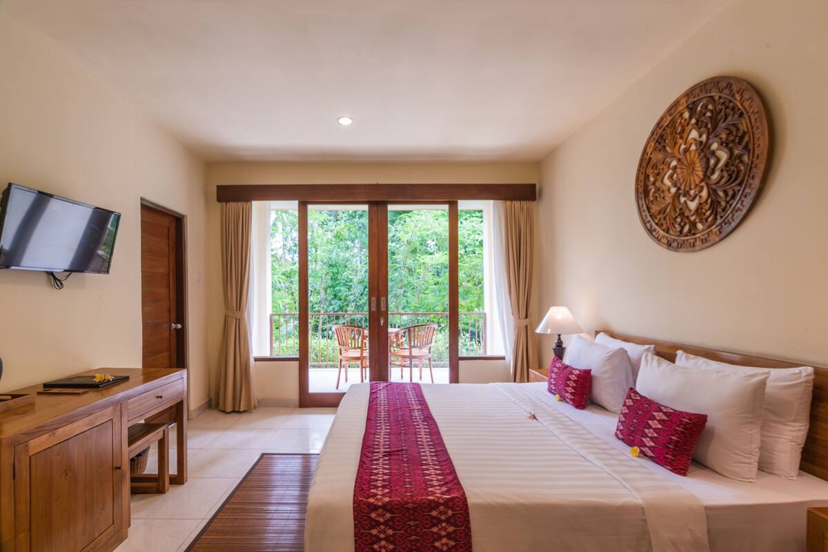 A hotel room with a bed, a TV, a wooden desk, a balcony with chairs, and a view of greenery.