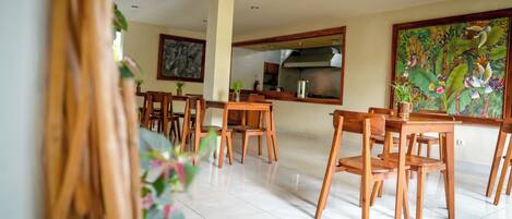 A dining area with wooden tables and chairs, a kitchen in the background, and a large floral painting on the wall.