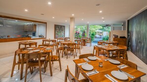 A spacious dining area with multiple wooden tables and chairs, a kitchen in the background, and a view of the outdoors.