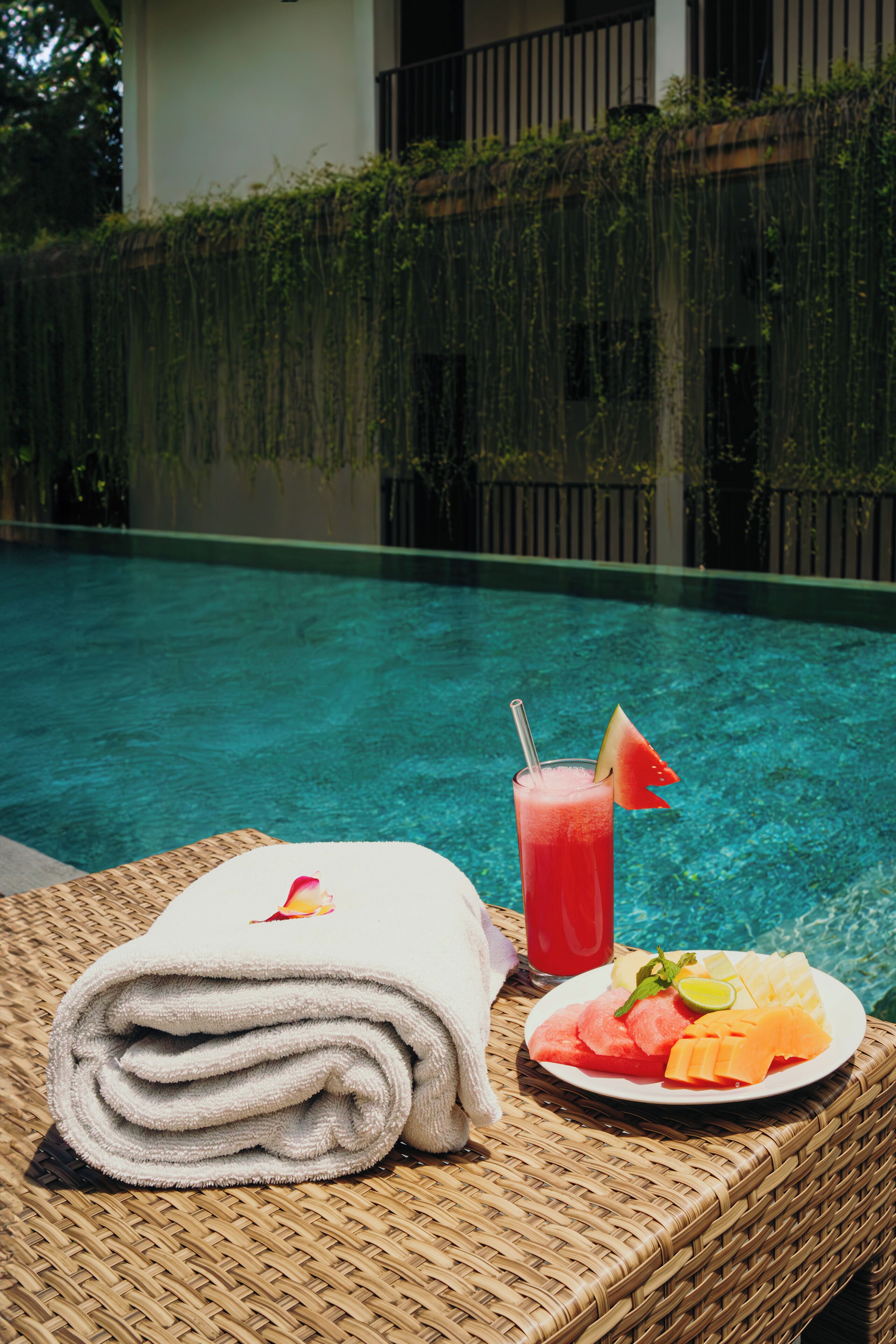 Une chaise en osier, une serviette pliée, une assiette de fruits et une boisson, le tout placé près d’une piscine.