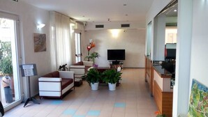 A hotel lobby with a sofa, television, plants, and a reception desk. - Hotel San Nazario (Varazze)