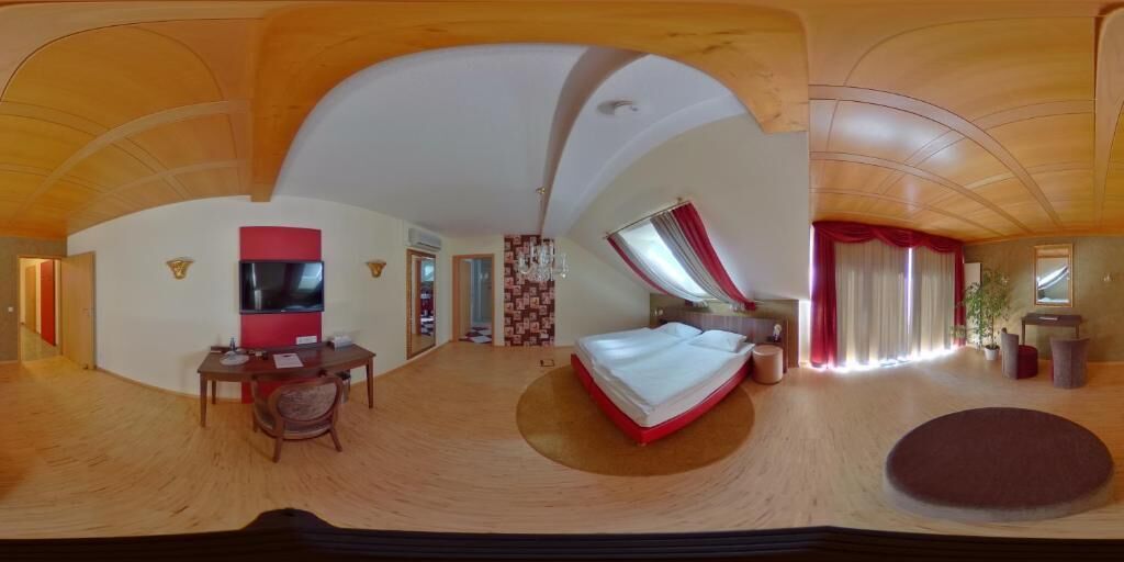 Ein weiter Blick auf ein Hotelzimmer mit gewölbter Decke, einem Bett, einem Schreibtisch mit Stuhl, einem Fernseher und einem großen Fenster mit Vorhängen.