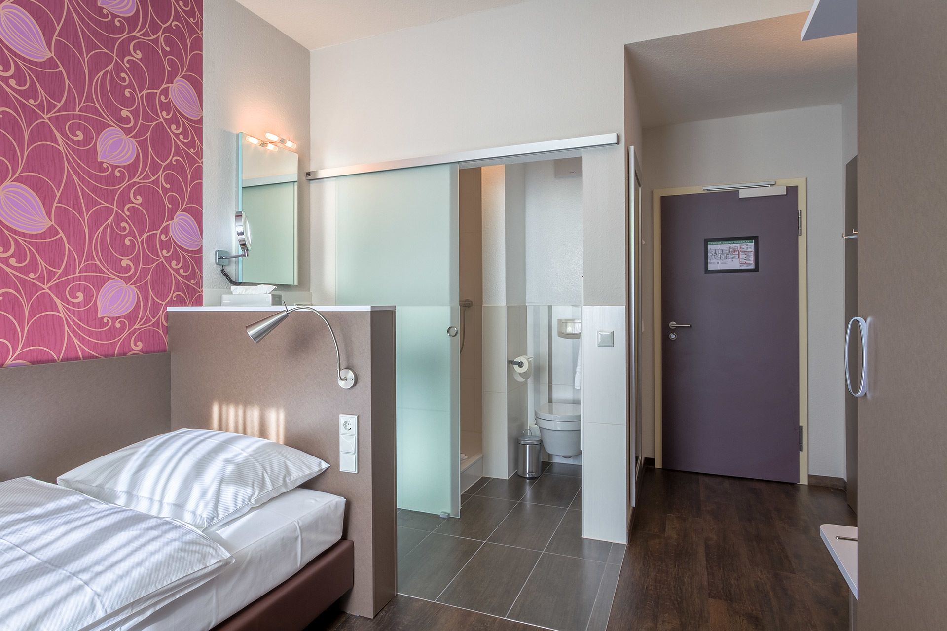 Hotelkamer met een bed, een badkamer met toilet en douche, en een deur naar een andere kamer.