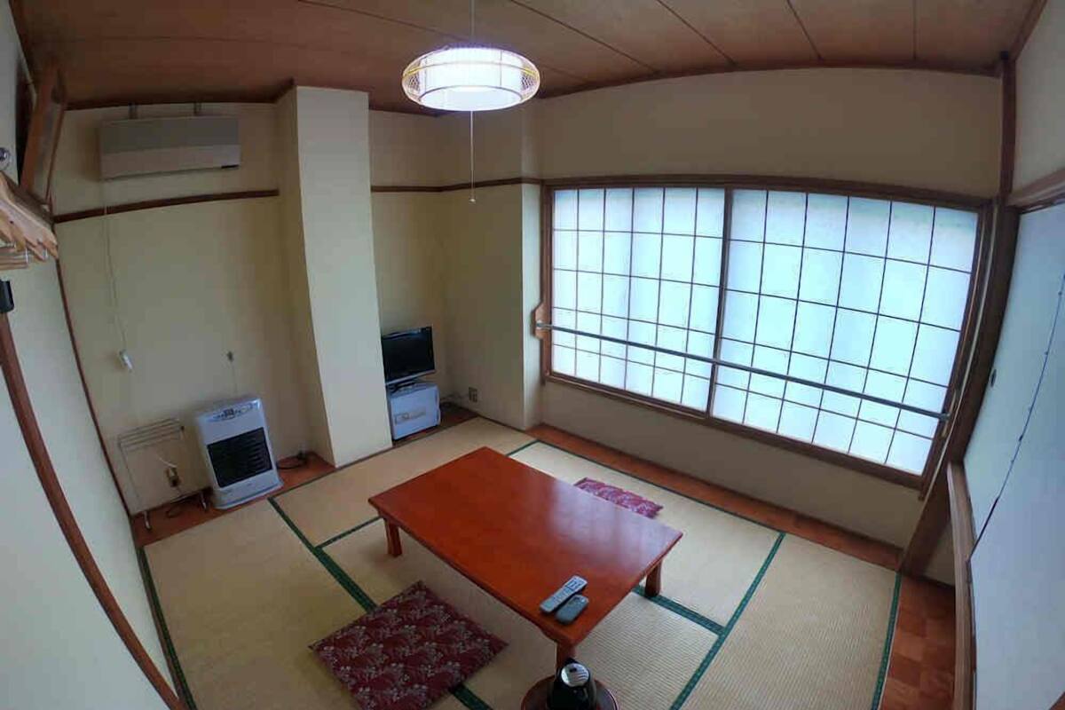 A traditional Japanese room with tatami flooring, a low table, a futon, a ceiling fan, and a large window with shoji screens.