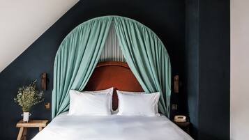 A bed with white linens, a wooden nightstand, a vase with flowers, and teal drapes.