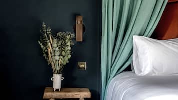 Un dormitorio con cama, un taburete de madera, un jarrón con flores, una lámpara de pared y un enchufe.