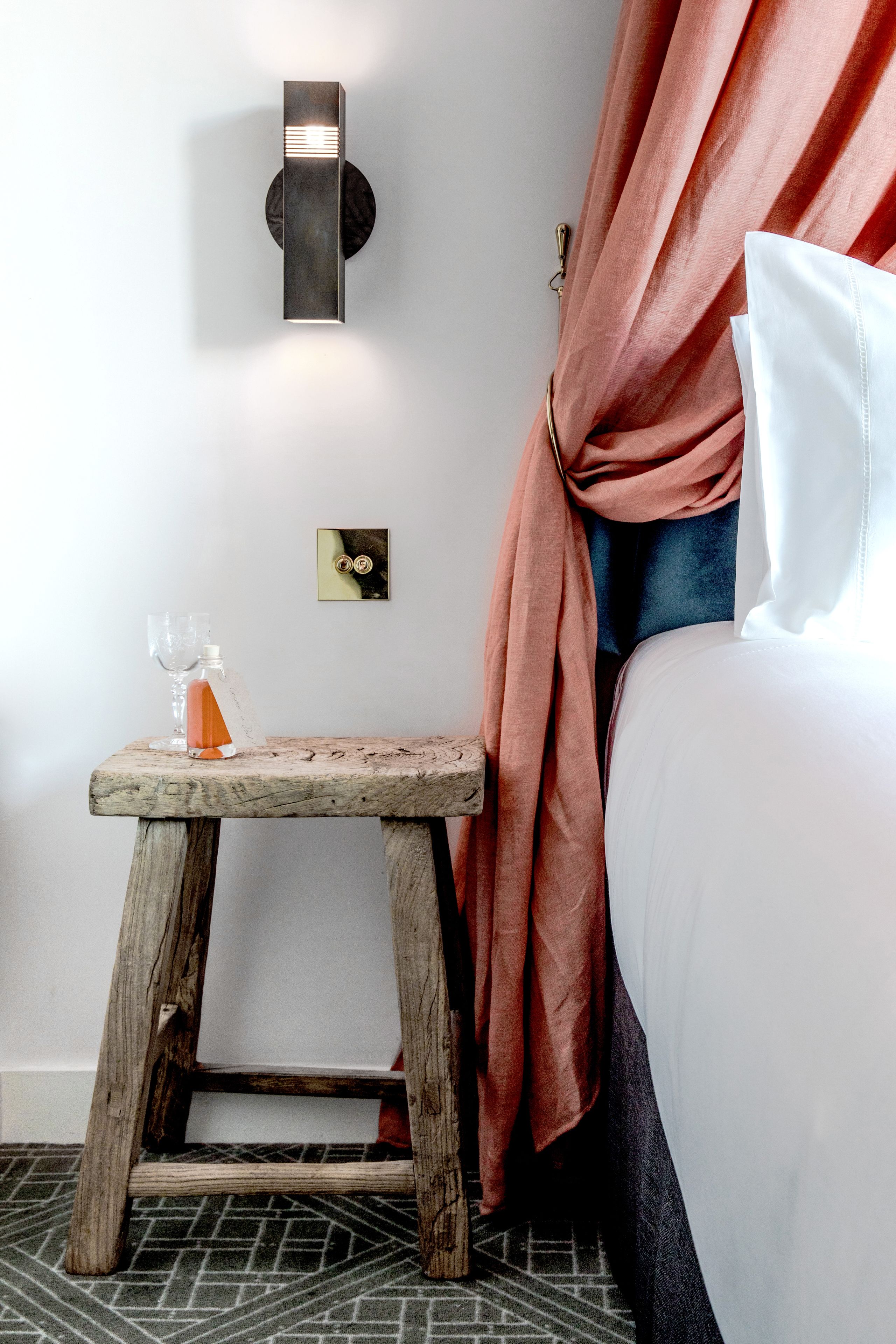 Un tabouret en bois, une bouteille et des verres, une lampe fixée au mur, un lit avec du linge de lit blanc et un rideau.