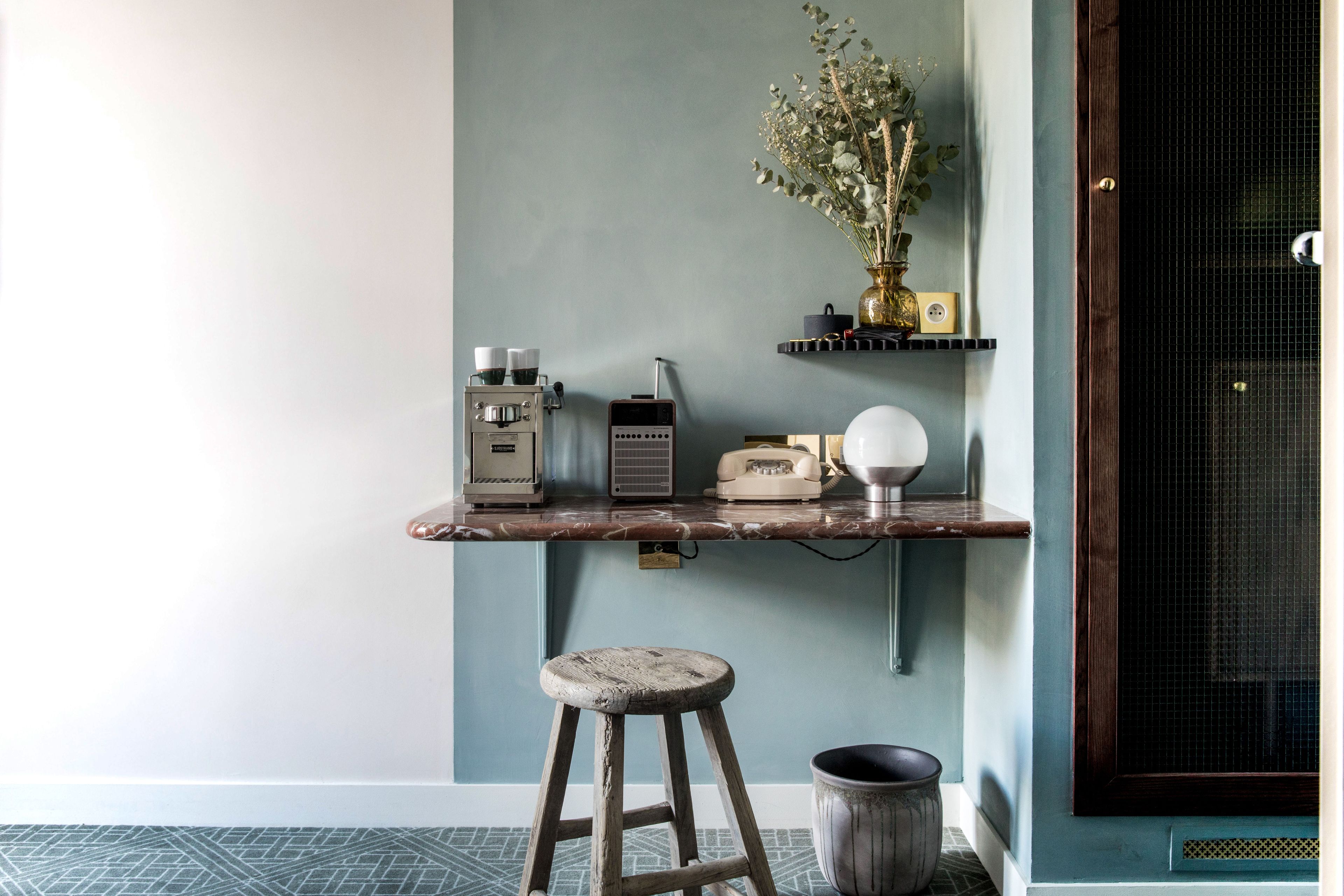 Una habitación con pared azul, un taburete de madera, un balde de metal, una radio antigua, un teléfono y un florero con flores.