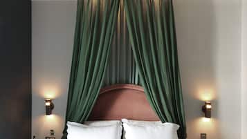 Cama com roupa de cama branca, cabeceira de madeira e cortinas verdes. Existem duas luzes fixadas na parede, de cada lado da cama.