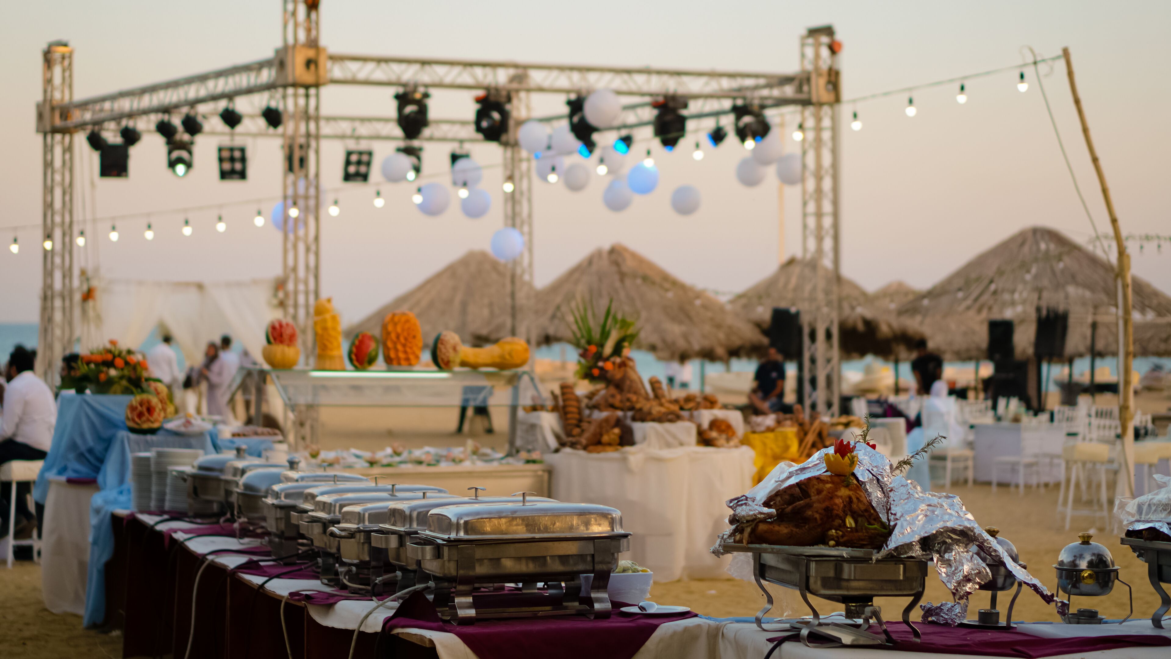 A buffet setup with food service equipment and tables, set up outdoors with thatched umbrellas in the background.