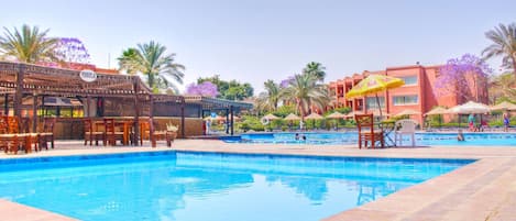 A swimming pool with lounge chairs and umbrellas, surrounded by palm trees and a building with a red facade.