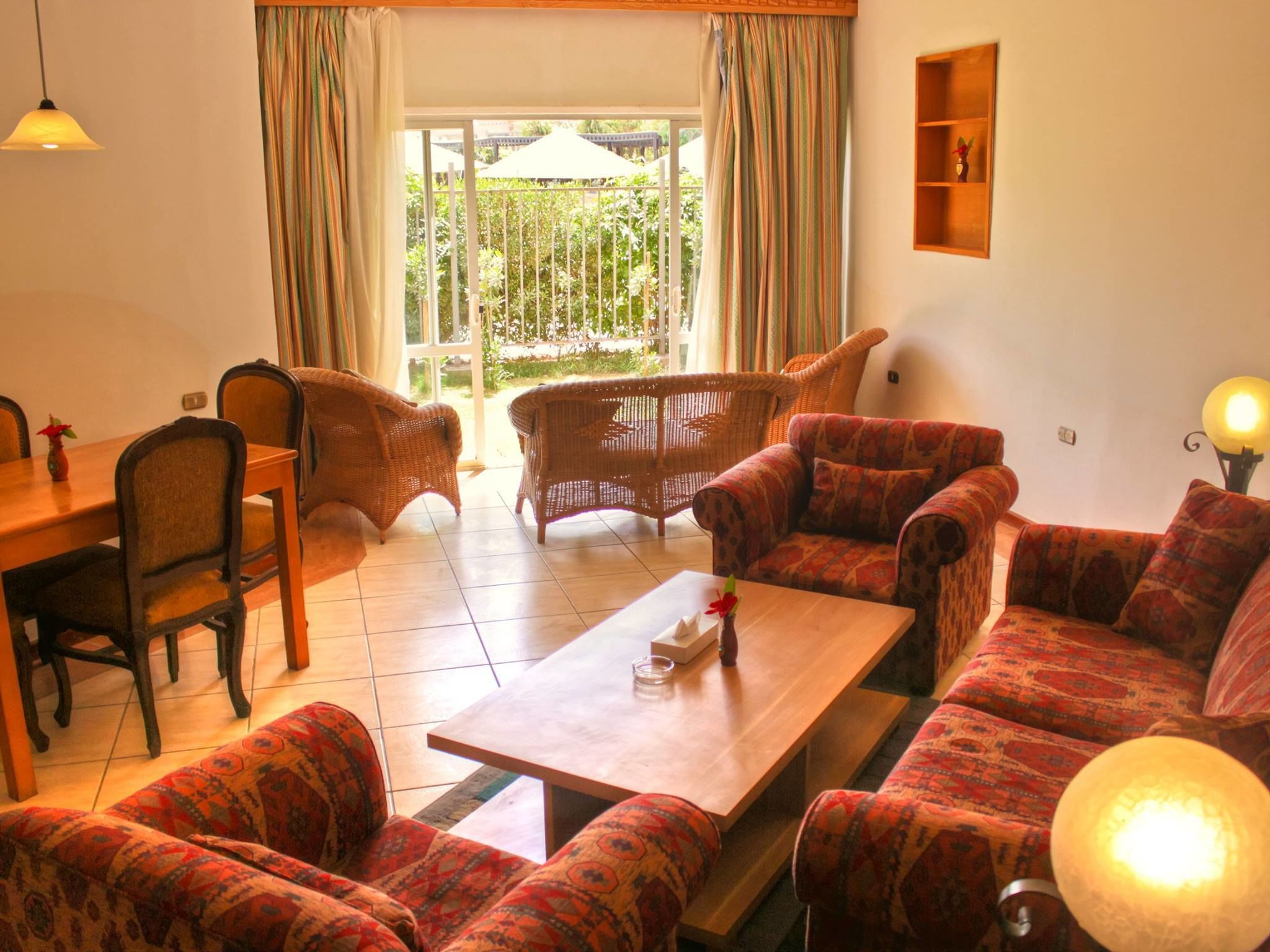 A living room with a dining table, two sofas, and a coffee table. There is a window with curtains, a shelf with decorative items, and a hanging lamp.