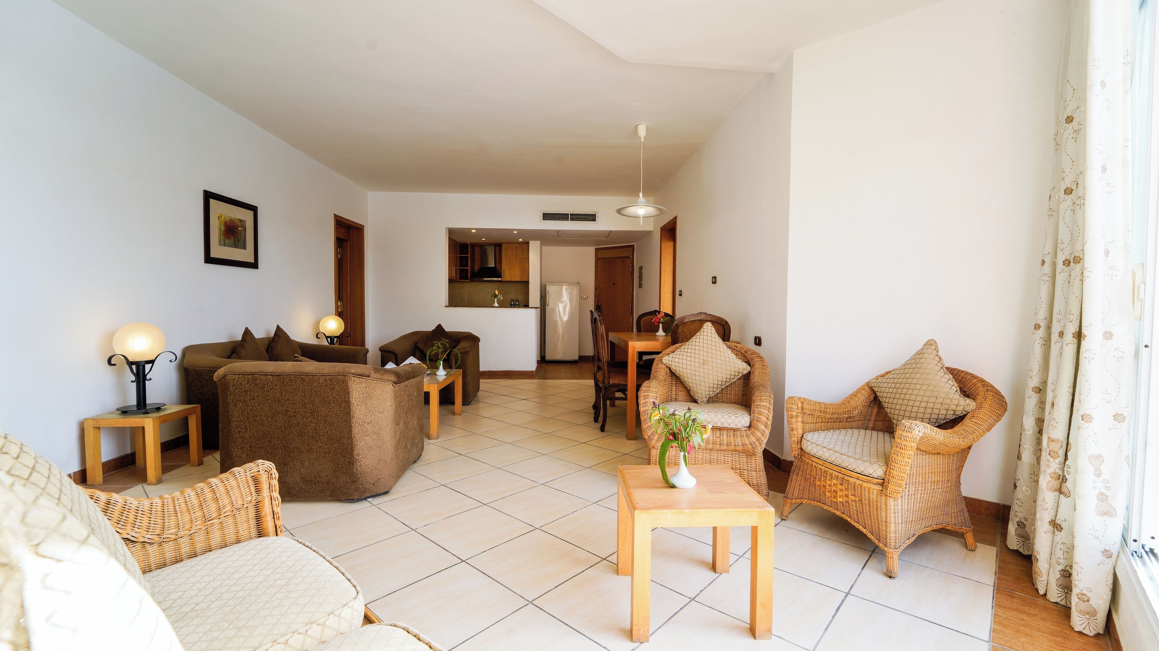 A living room with a sofa, two armchairs, a coffee table, and a dining table. There is a kitchen in the background with a refrigerator and a microwave.