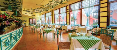 A restaurant with multiple tables and chairs, large windows with curtains, and hanging pendant lights.